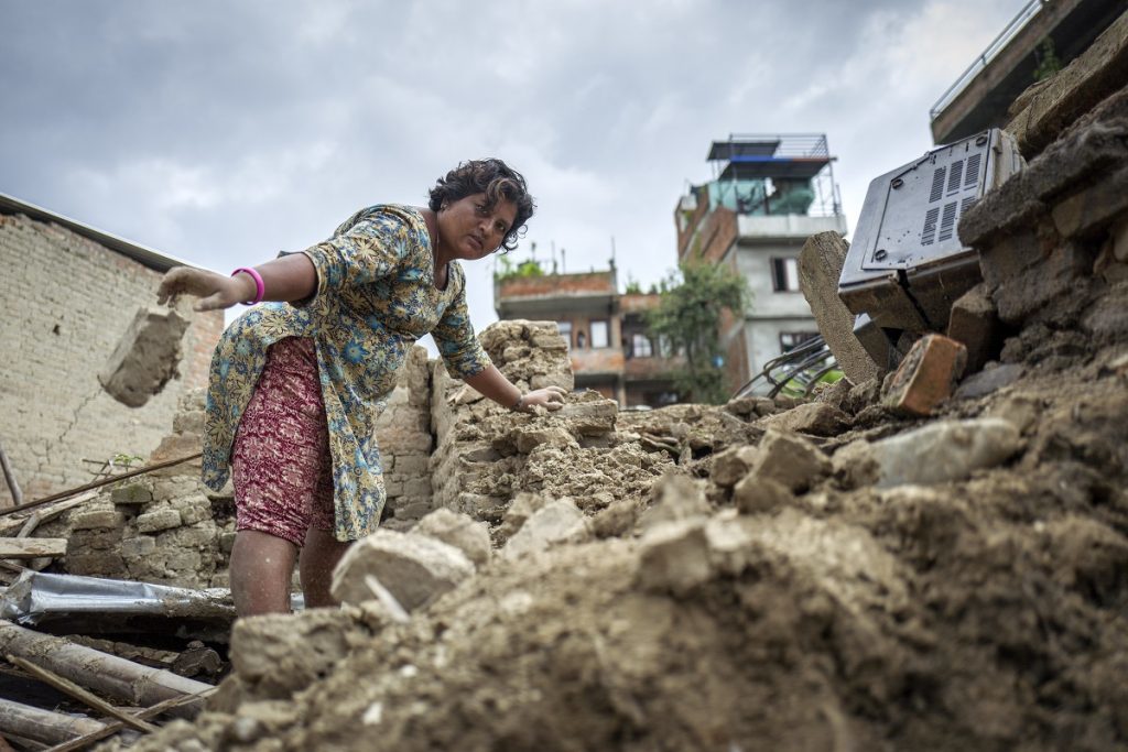A Nepalese woman stands atop rubble from the 2015 Nepal earthquakes and throws a brick towards the ground. Women's leadership is at the center of ActionAid's emergency response.