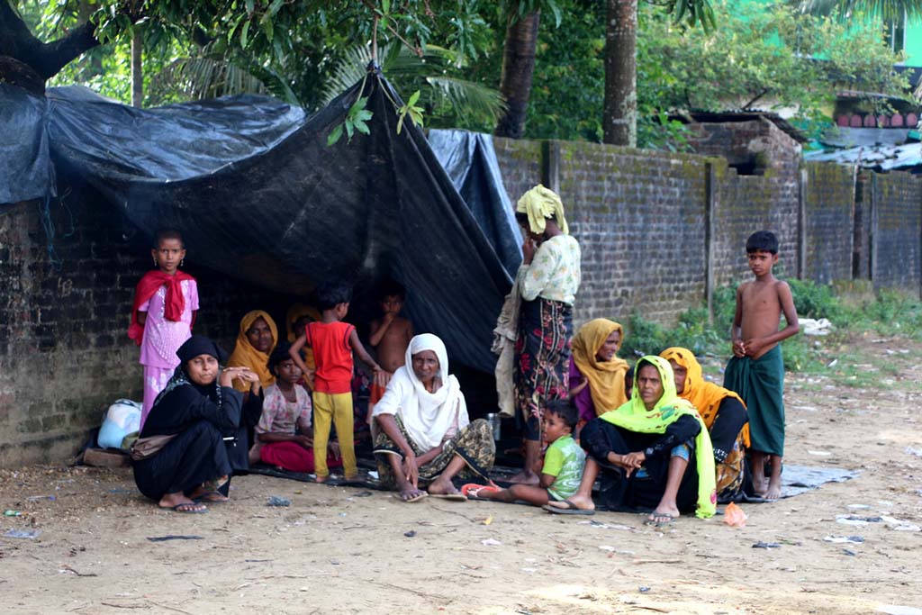 A group of women and children sit under a makeshift tent by the road.