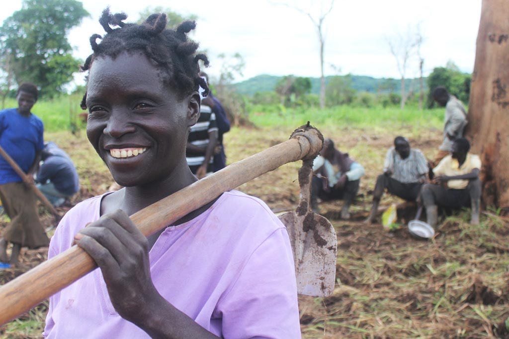 A woman farmer from Uganda is smiling and resting a garden hoe on her shoulder.