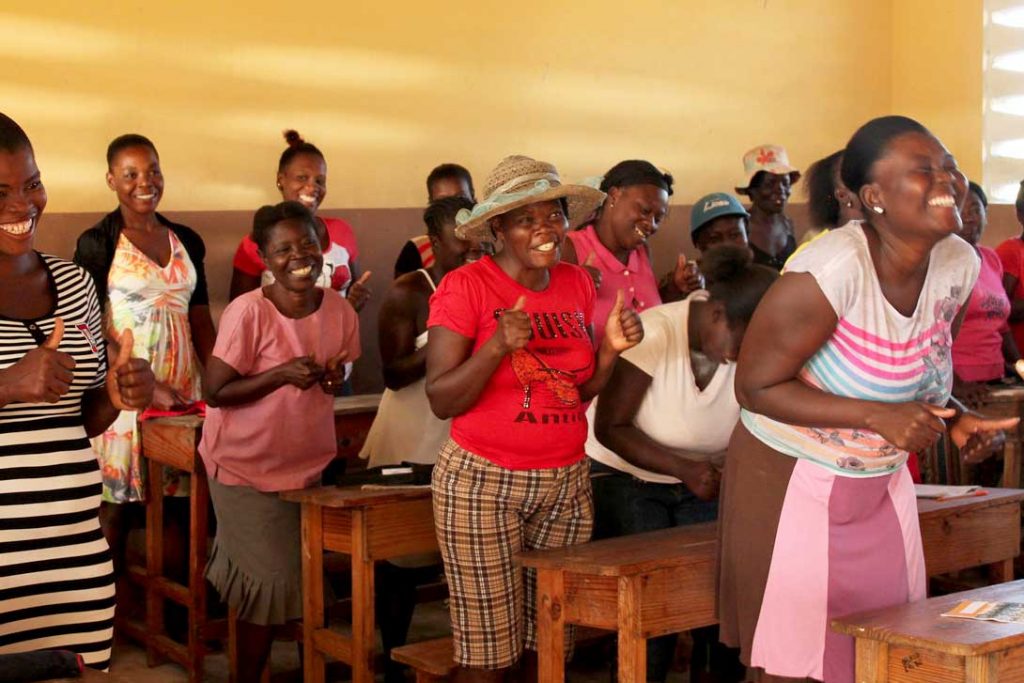 Women stand behind desks in rows with their thumbs up as part of a protection training in Haiti after Hurricane Matthew.