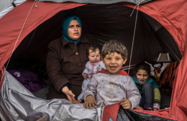 A Syrian refugee woman sits in a tent with her three children. Her young son is standing the foreground, smiling.