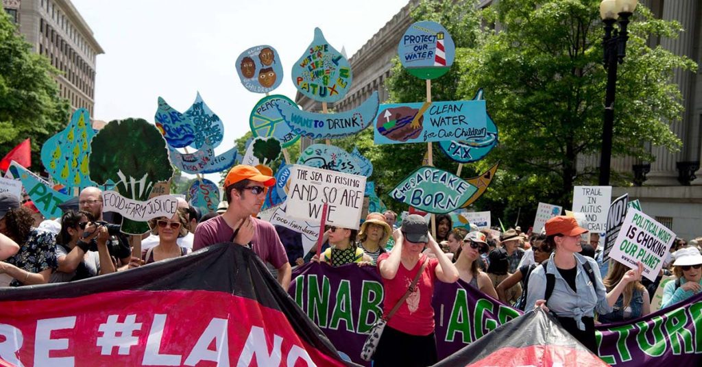 A group of people stand and hold up signs with text that call for protecting water and defending food and farm justice - things that Sam Clovis wouldn't do.