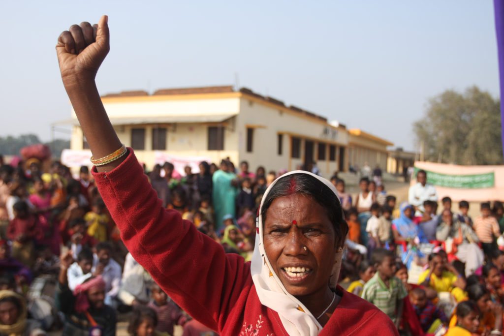 A Dalit woman in India with a determined look on her face raises her right fist. In the background is a crowd of people. They participated in a land rights march that included women farmers.