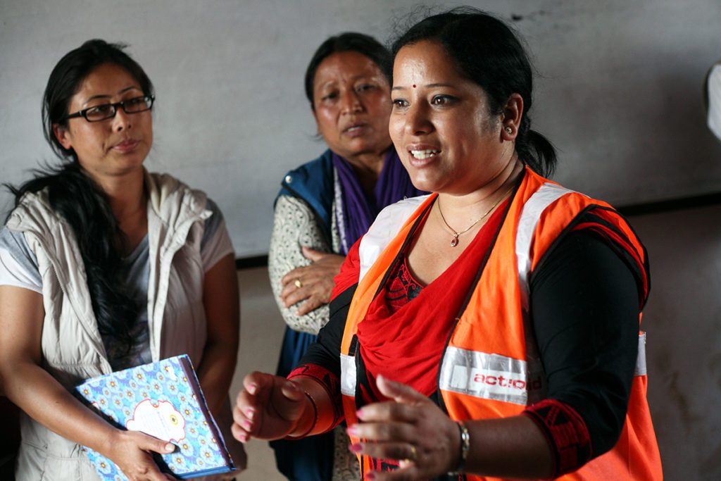 A woman in an orange vest addresses a group (not pictured), while two women stand in the background, listening.