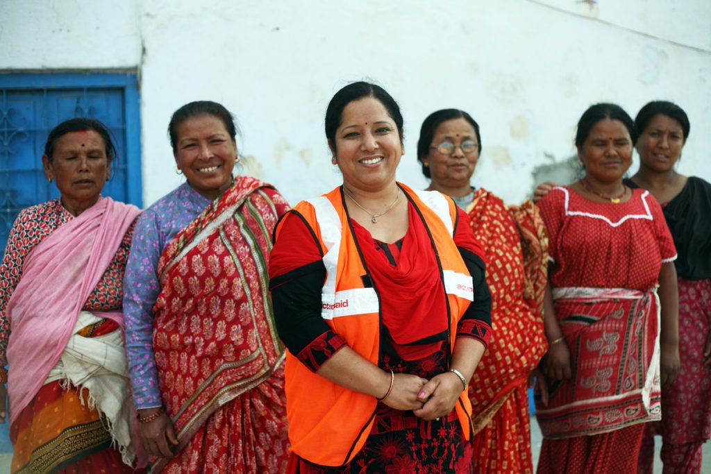 Six women stand against a white wall. A woman in an orange vest bearing ActionAid's 