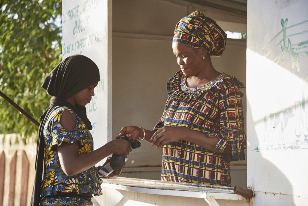 A woman standing at the window of a small structure hands a small black bag over the counter to a girl.