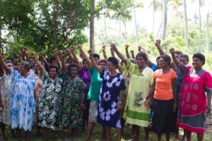 About 30 women in brightly colored dresses are smiling and standing in rows with their right fists raised.