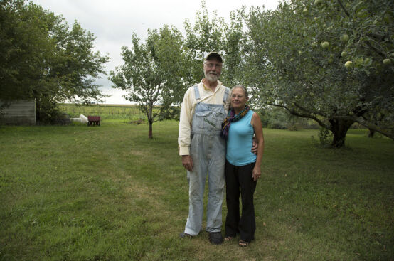 A man and a woman stand next to each other on a grassy field. The man, dressed in overalls, has his left arm around the woman.