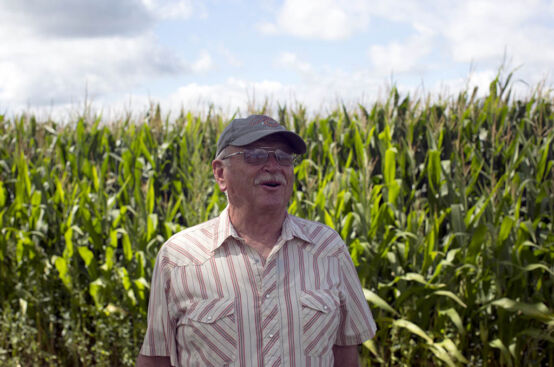 A man with a white mustache and a cap stands in front of cornstalks.